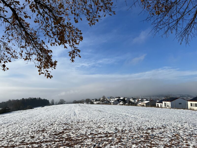 Panoramablick über Pfaffenhofen. Im Vordergrund ein weitgehend verschneites Feld ein paar Häuser, hereinhängende Zweige von einem Baum und weitgehend blauer Himmel. Im Hintergrund eine Dunstglocke, der Kirchturm ist kaum zu erkennen und die Hügel dahinter gar nicht.