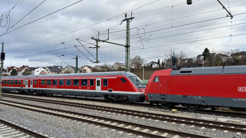 Auf dem 4. Gleis vom Betrachter aus fährt ein Regionalzug aus dem Bahnhof, zu sehen sind ein halber Mittelwagen - mit gelben Aufklebern auf einer Tür - und ein Steuerwagen in frischem, kräftigen Rot mit weißem Streifen, beschriftet mit 'München-Nürnberg-Express' und einem großen Fahrradsymbol im Fenster, dahinter zur
Hälfte die rote Lok, Baureihe 101.