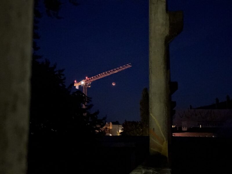 Der verfinsterte rote Mond unter einem beleuchteten Baukran, fotografiert durch graue Betonsäulen hindurch.