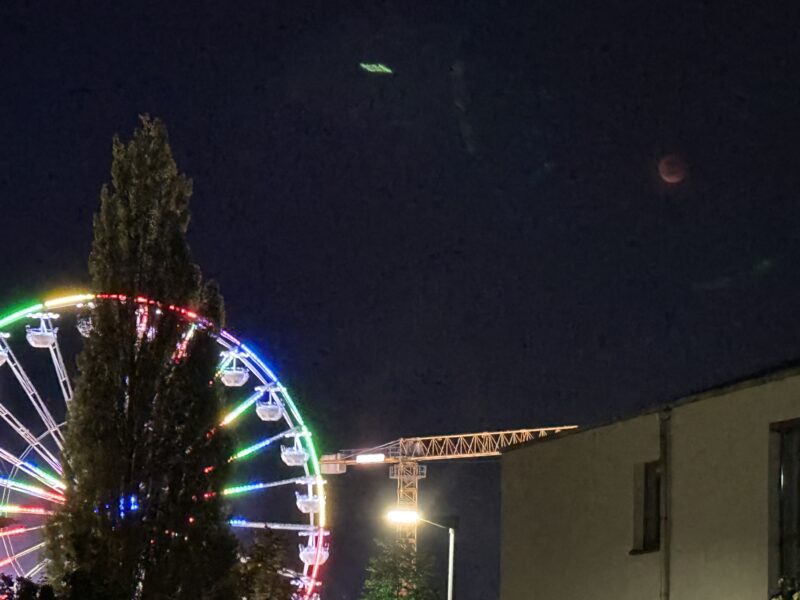 Links ein regenbogenbunt beleuchtetes Riesenrad hinter einem hohen Baum, in der Mitte ein beleuchteter Baukran, rechts unten ein Haus und oben der rote verfinsterte Mond, relativ blass. Linsenreflexionen vom Riesenrad sind auch am Himmel zu sehen.