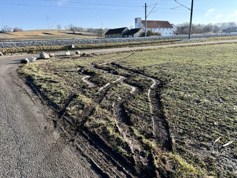 Eck eines Feldes, auf dem ein paar krumme, überlagerte Furchen, die offenbar von einem Auto stammen, zu sehen sind. Das Eck des links und hinten verlaufenden asphaltierten Weges ist mit ein paar großen Steinen versehen. Im Hintergrund eine Bahnlinie und landwirtschaftliche Gebäude.