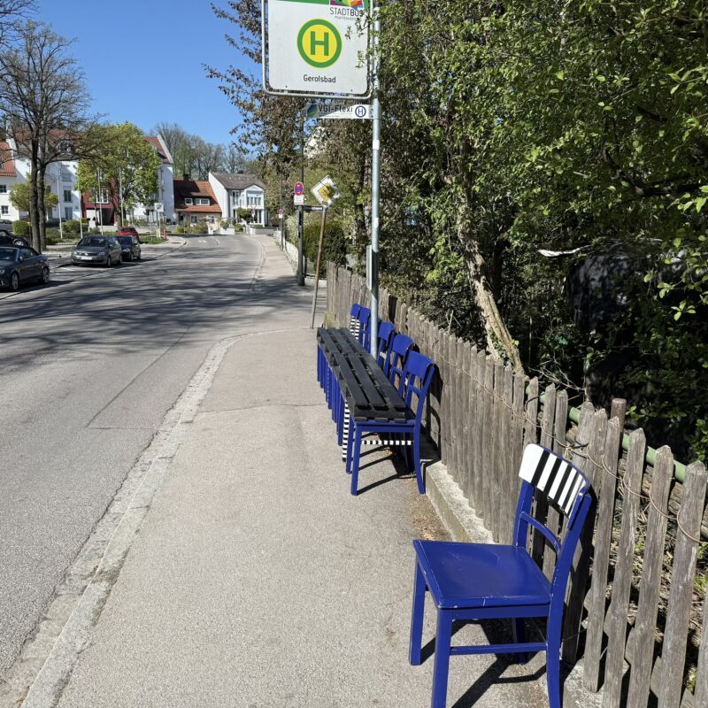 Gehweg mit Bushaltestelle "Gerolsbad", links die Fahrbahn, rechts ein Gartenzaun aus alten graubraunen Holzlatten, dahinter viel Grün. Kein Wartehäuschen o.ä., aber eine Gruppe von 6 einfachen Stühlen in einem kräftigen mitteldunklen Blau, wobei einzelne Stangen oder Beine in einem schwarz-weißen Streifenmuster gestrichen sind. Quer über die Stühle liegen 2 Teile einer schwarzen Holzpalette (je 3 Bretter breit), die somit die Sitzfläche bilden. Alles noch recht neu und sauber. Mit ca. 2 Metern Abstand steht im Bildvordergrund ein siebter, einzelner Stuhl ohne Palette; bei diesem ist es die Rückenlehne, die schwarz-weiß gestreift ist.