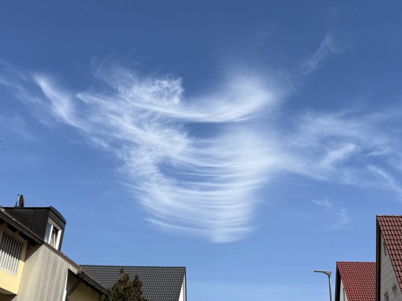 Blauer Himmel mit einer dünnen weißen Wolkenstrukrur: rechts etwas dicker, in der Mitte unregelmäßige horizontale Linien, links wie Flaumfedern verwirbelt. Darunter ein paar Hausdächer.