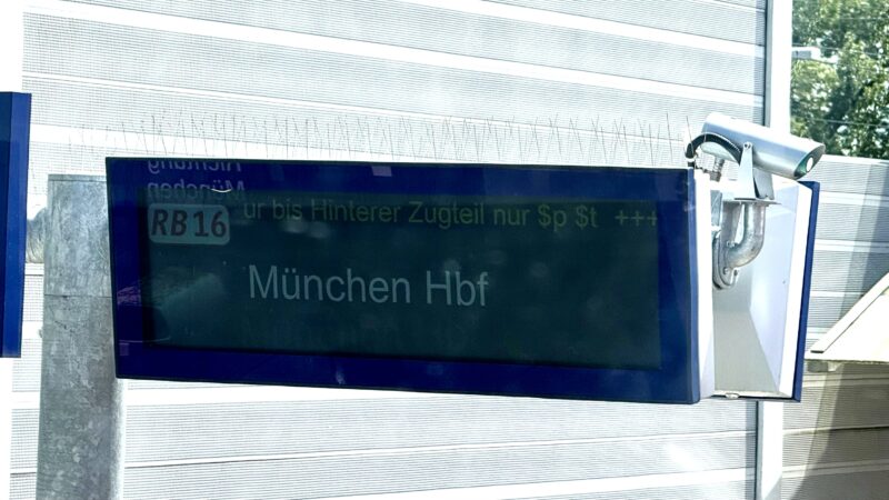 Anzeigebildschirm am Bahnsteig, aus dem Zug heraus mit nicht ganz sauberer Scheibe und ungünstigen Lichtverhältnissen fotografiert, im Hintergrund v.a. Schallschutzwände. Auf der Anzeige: RB 16 München Hbf, die Laufschrift zeigt gerade 'ur bis Hinterer Zugteil nur $p $t +++'
